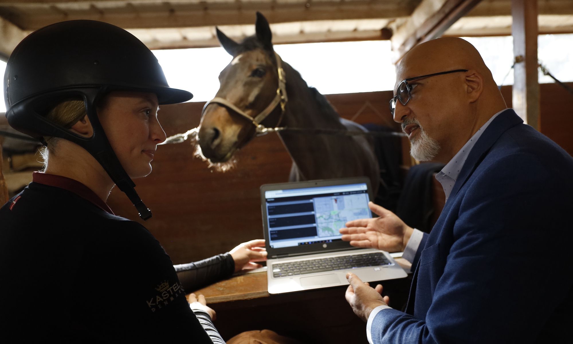 An equestrian in a helmet listens to a professional showing data on a laptop, with a horse in a stable observing nearby, highlighting collaboration between technology and equestrian care